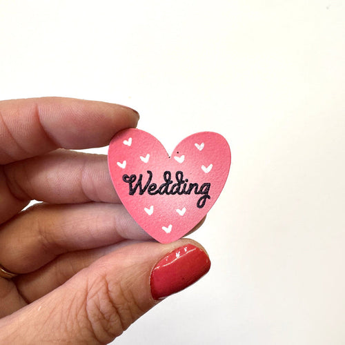 A hand holds a small pink heart-shaped magnet with white heart details and the word 'Wedding' written in black script against a plain white background.