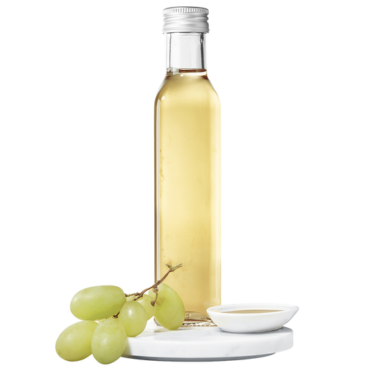 A clear bottle of white balsamic vinegar with a silver cap, accompanied by a small white dish and a bunch of green grapes.