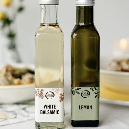 a White balsamic bottle and Lemon Olive oil bottle next to each other on a white table with dishes and food blurred in the background