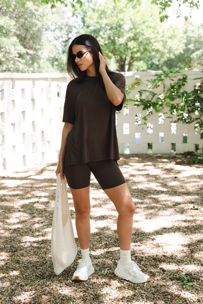 Female posing standing outside in front of a white fence wearing the Women’s Biker Short Set in Espresso