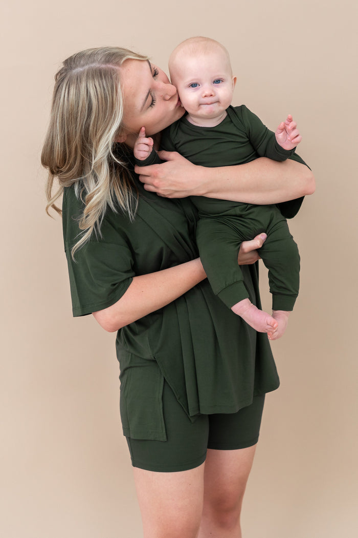 Woman holding a baby both wearing green outfits against a beige background