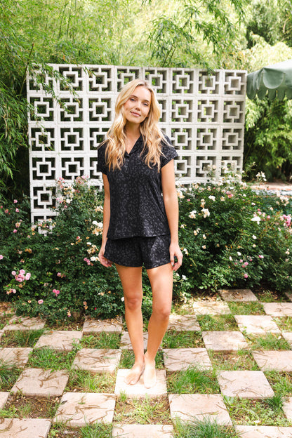 Female posing outside in front of a flower bush and white lattice wearing the Women’s Short Sleeve Pajama Set in Midnight Leopard